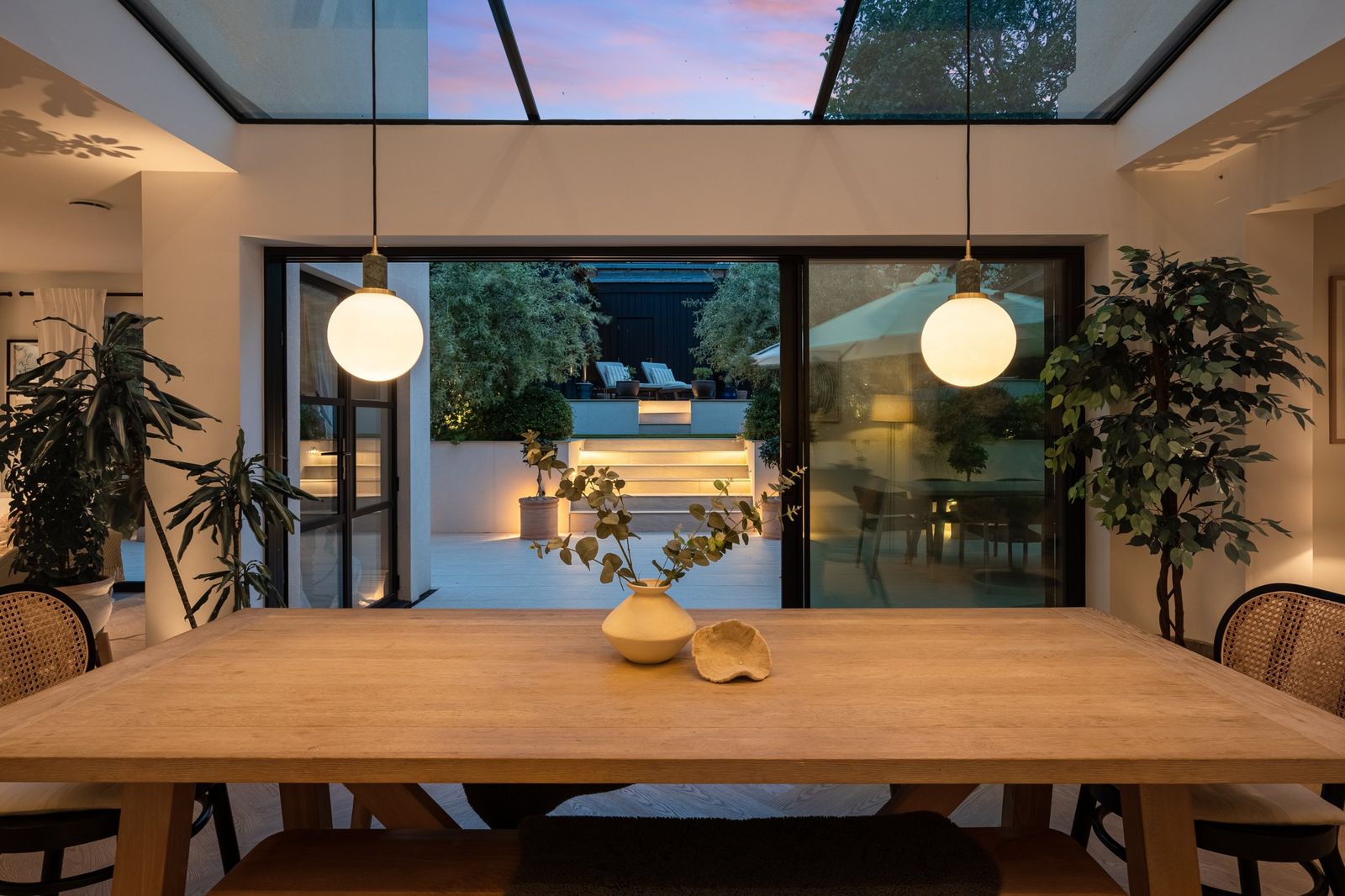The dining table under the roof glass at dusk, with the lit garden beyond