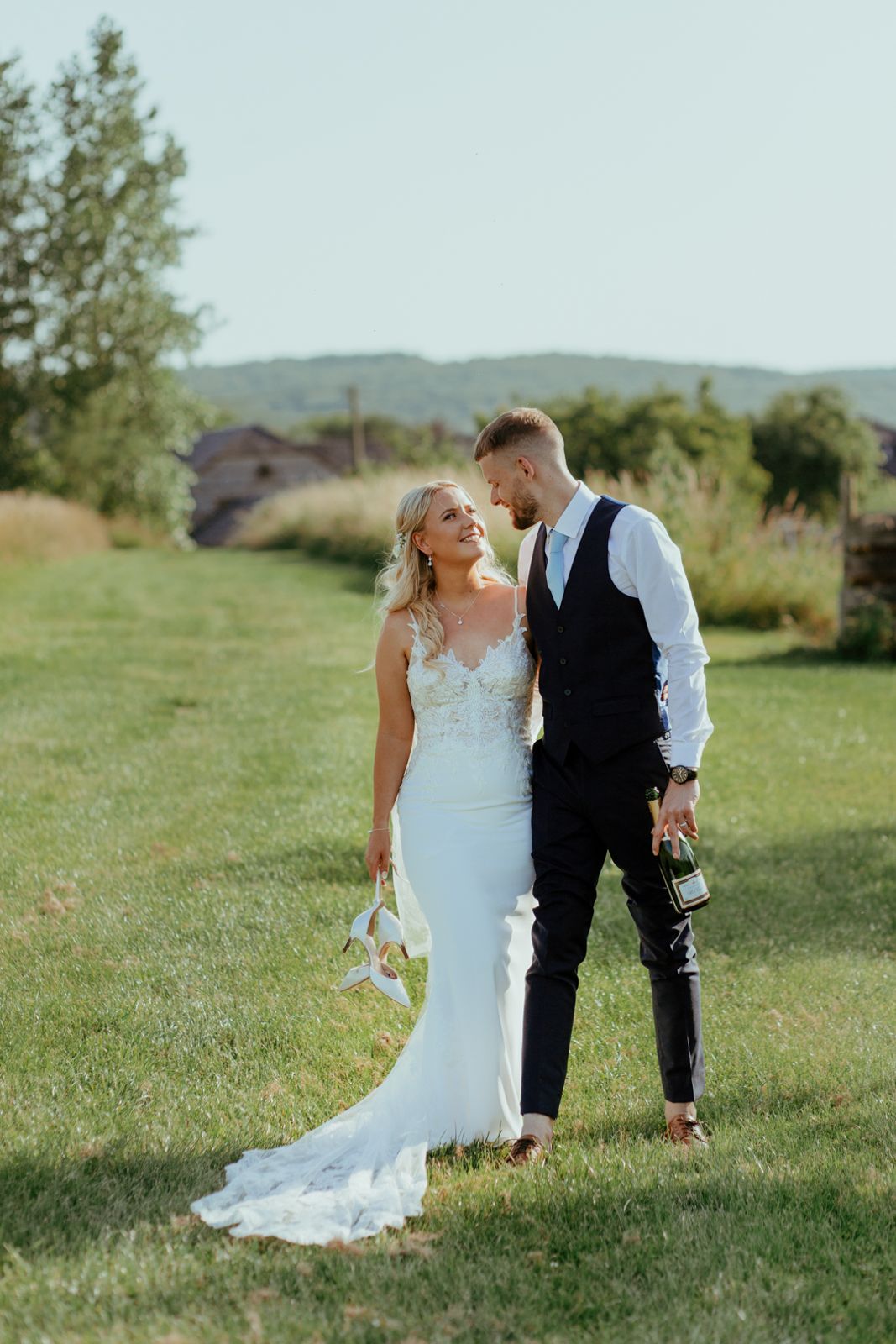 The couple walking across the grass with her shoes and a bottle of fizz